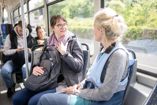 Zwei Personen unterhalten sich in der Straßenbahn. Eine Person ist mit einer blauen Weste als Diskursbegleiter:in von Platz nehmen für Demokratie gekennzeichnet.
