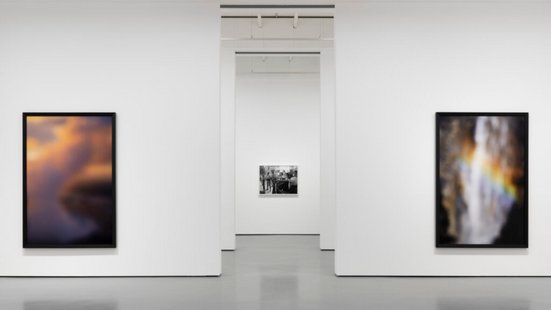 Hochformatiges Sharepic in einem modernen, wei&szlig;en Ausstellungsraum: An der linken Wand h&auml;ngt ein gerahmtes, unscharf wirkendes Foto in warmen Farben wie Orange, Violett und Dunkelblau