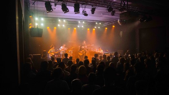 Mehrere Musiker*innen mit Schlagzeug, E‑Gitarre, Bass, Keyboard und einer Person mit Akustikgitarre im Zentrum am Mikrofon auf der B&uuml;hne. Orange und wei&szlig;e Scheinwerfer leuchten durch Nebel, vorn die Silhouetten eines dicht gedr&auml;ngten Publikums.