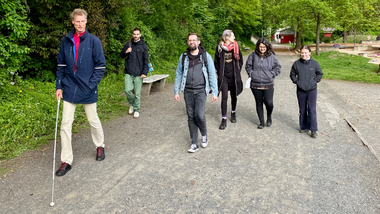 Sechs Personen gehen durch eine grüne Parkanlage. Vorne links eine Person mit weißem Langstock in dunkelblauer Jacke und beiger Hose, dahinter fünf weitere Personen in Freizeitkleidung. Üppiges Grün säumt den asphaltierten Weg.