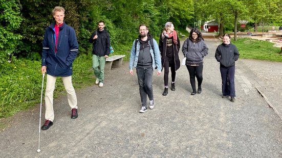 Sechs Personen gehen durch eine gr&uuml;ne Parkanlage. Vorne links eine Person mit wei&szlig;em Langstock in dunkelblauer Jacke und beiger Hose, dahinter f&uuml;nf weitere Personen in Freizeitkleidung. &Uuml;ppiges Gr&uuml;n s&auml;umt den asphaltierten Weg.