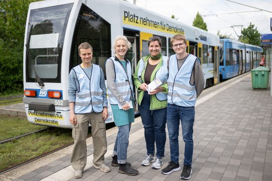 Vier Personen stehen als Diskursbegleiter:innen mit blauen Westen gekennzeichnet, vor dem Demokratiezug, einem Wagen der Stra&szlig;enbahnlinie 1. 